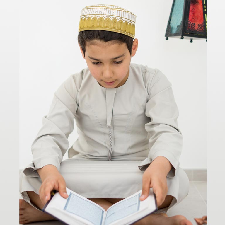 A young Muslim boy wearing a traditional Islamic cap and white attire, sitting on the floor and reciting the Quran with deep concentration. He is engaged in Quran memorization (Hifz), symbolizing dedication and spiritual growth.